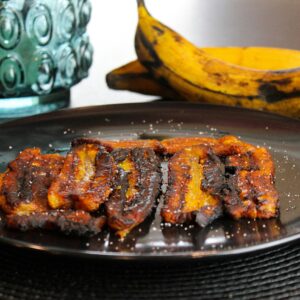 Fried plantains served on a black plate with fresh bananas in the background.